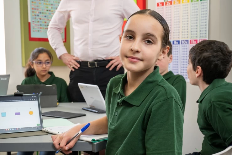 Image shows young female student pensively looking at the camera. There is a laptop showing a test question behind her, as well as her classmates and teacher.