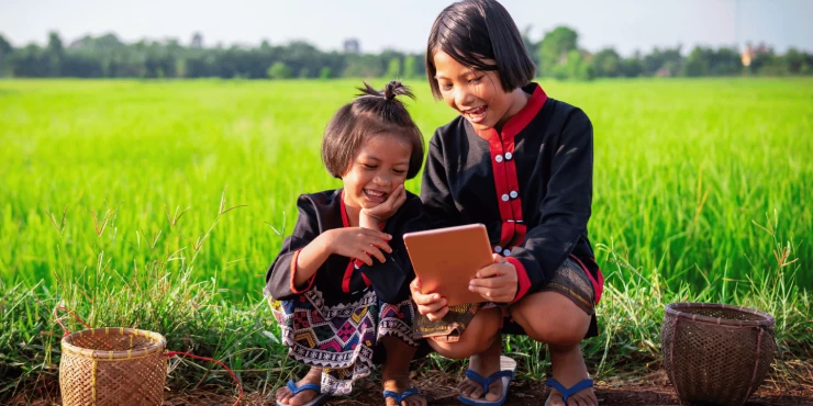 Two girls are crouched down looking at a tablet in a field in Asia. They are wearing traditional dresses.
